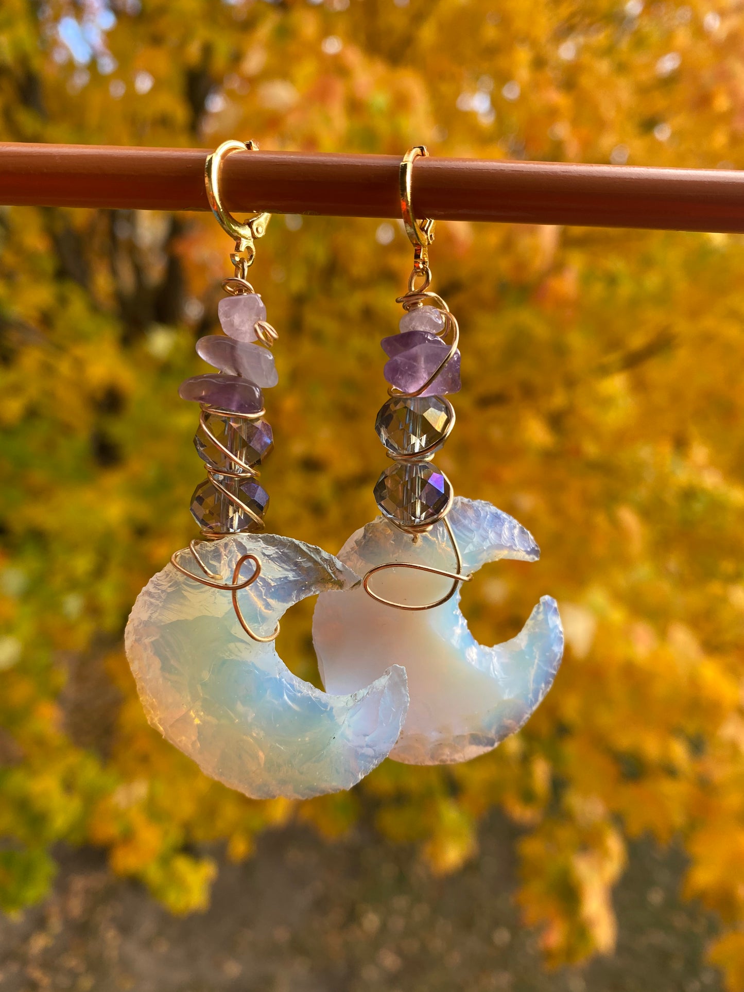 Amethyst and violet iridescent crystal beads with multicolored opalite moons, wrapped with gold wire to create dangle earrings. They are held onto a brown display by gold huggy closures. There is a gold tree and green landscape in the background.