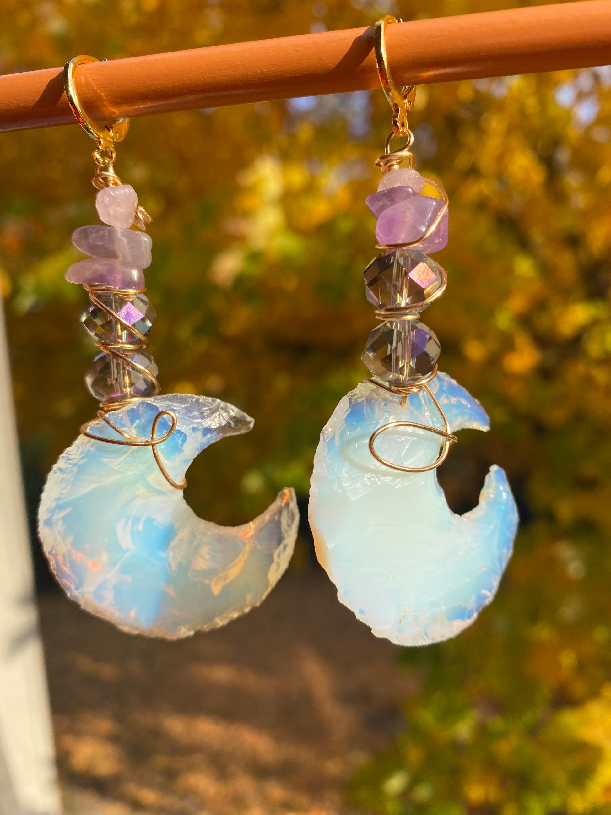 Amethyst and violet iridescent crystal beads with multicolored white opalite moons, wrapped with gold wire to create dangle earrings. They are held onto a brown display by gold huggy closures. There is a gold tree and green landscape in the background. 