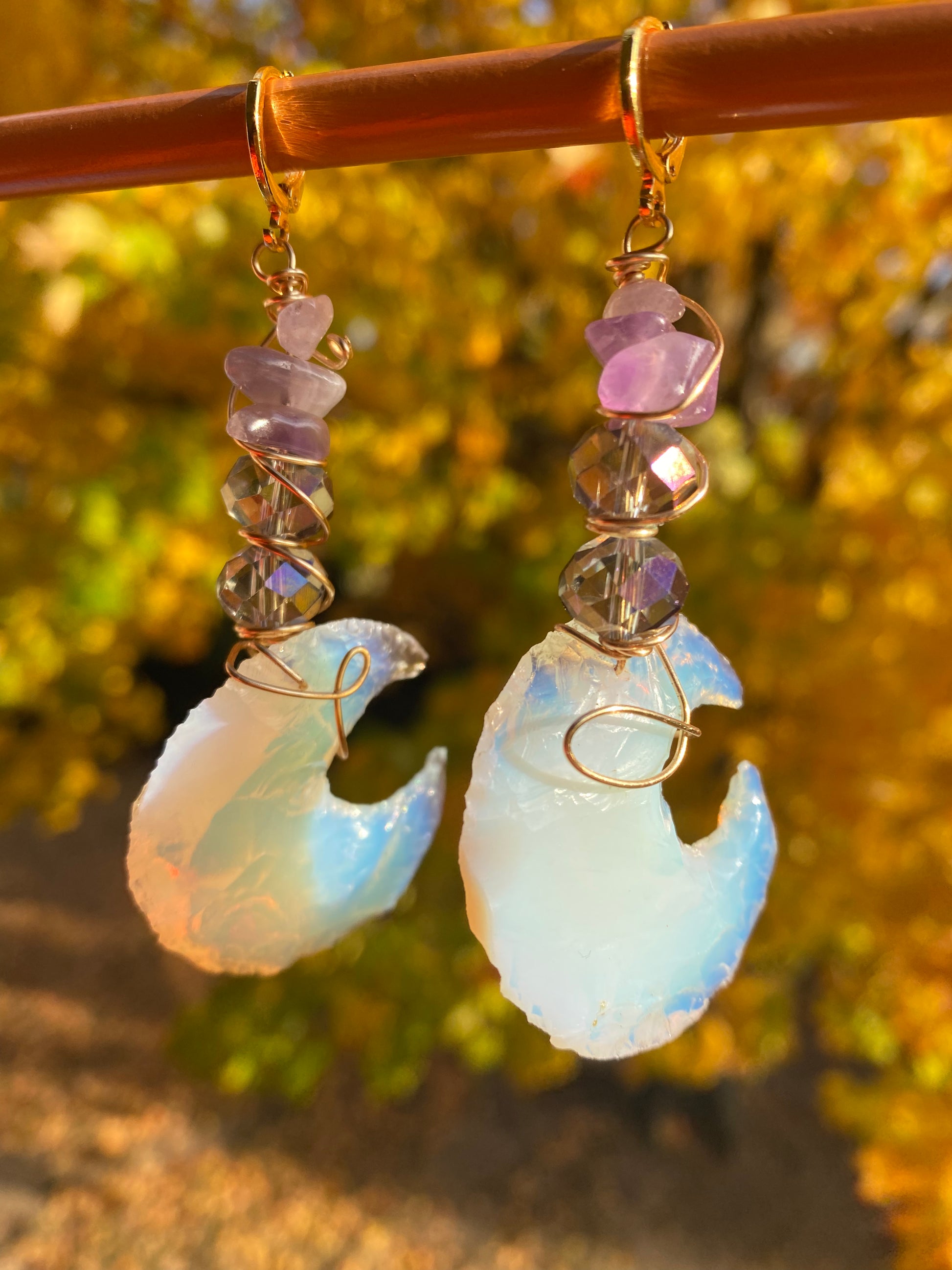 Amethyst and violet iridescent crystal beads with multicolored opalite moons, wrapped with gold wire to create dangle earrings. They are held onto a brown display by gold huggy closures. There is a gold tree in the background. Light bounces off of the earrings.