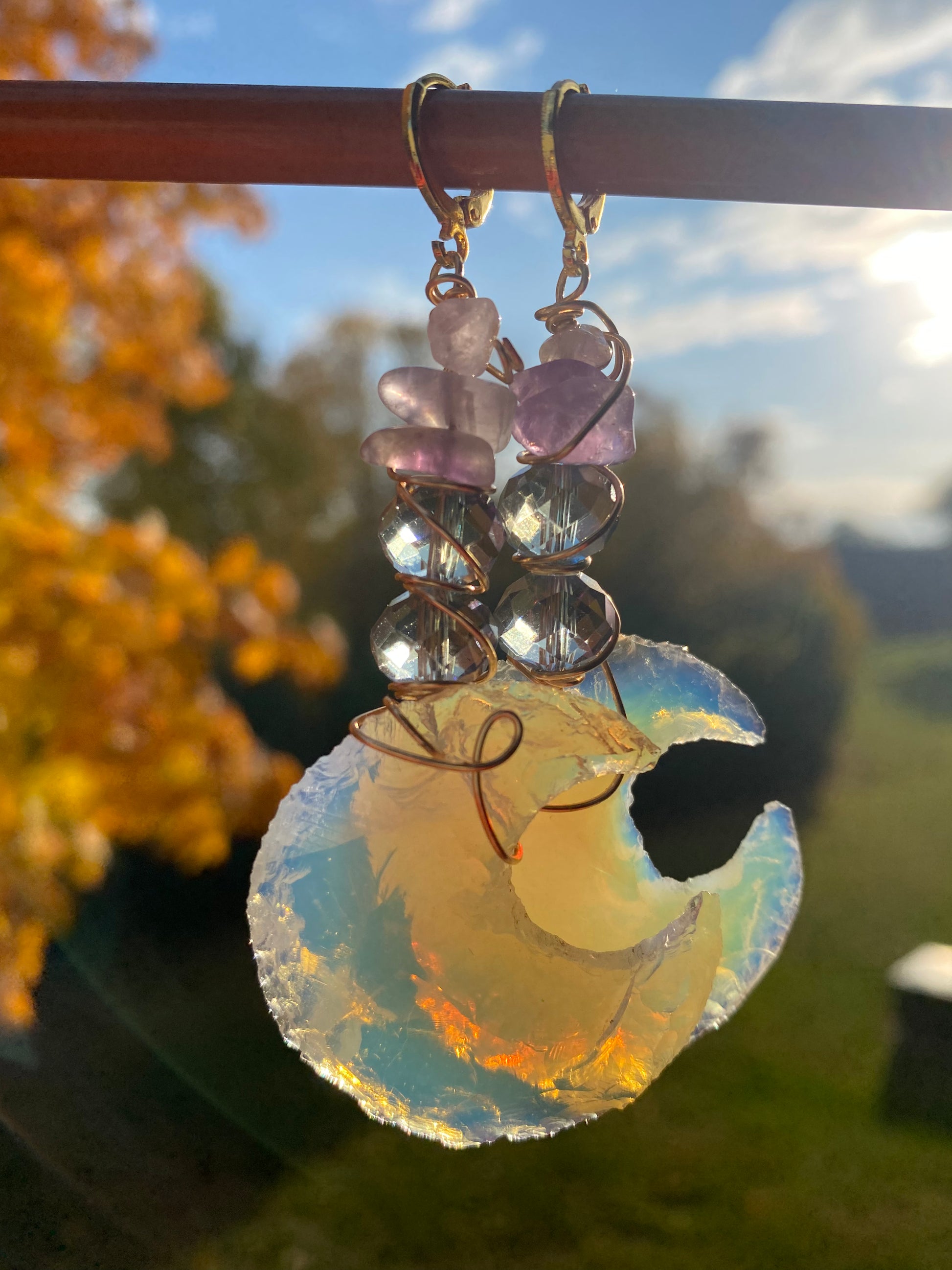 Amethyst and violet iridescent crystal beads with multicolored opalite moons, wrapped with gold wire to create dangle earrings. They are held onto a brown display by gold huggy closures. There is a gold tree, Blue sky, white clouds and green landscape in the background. 