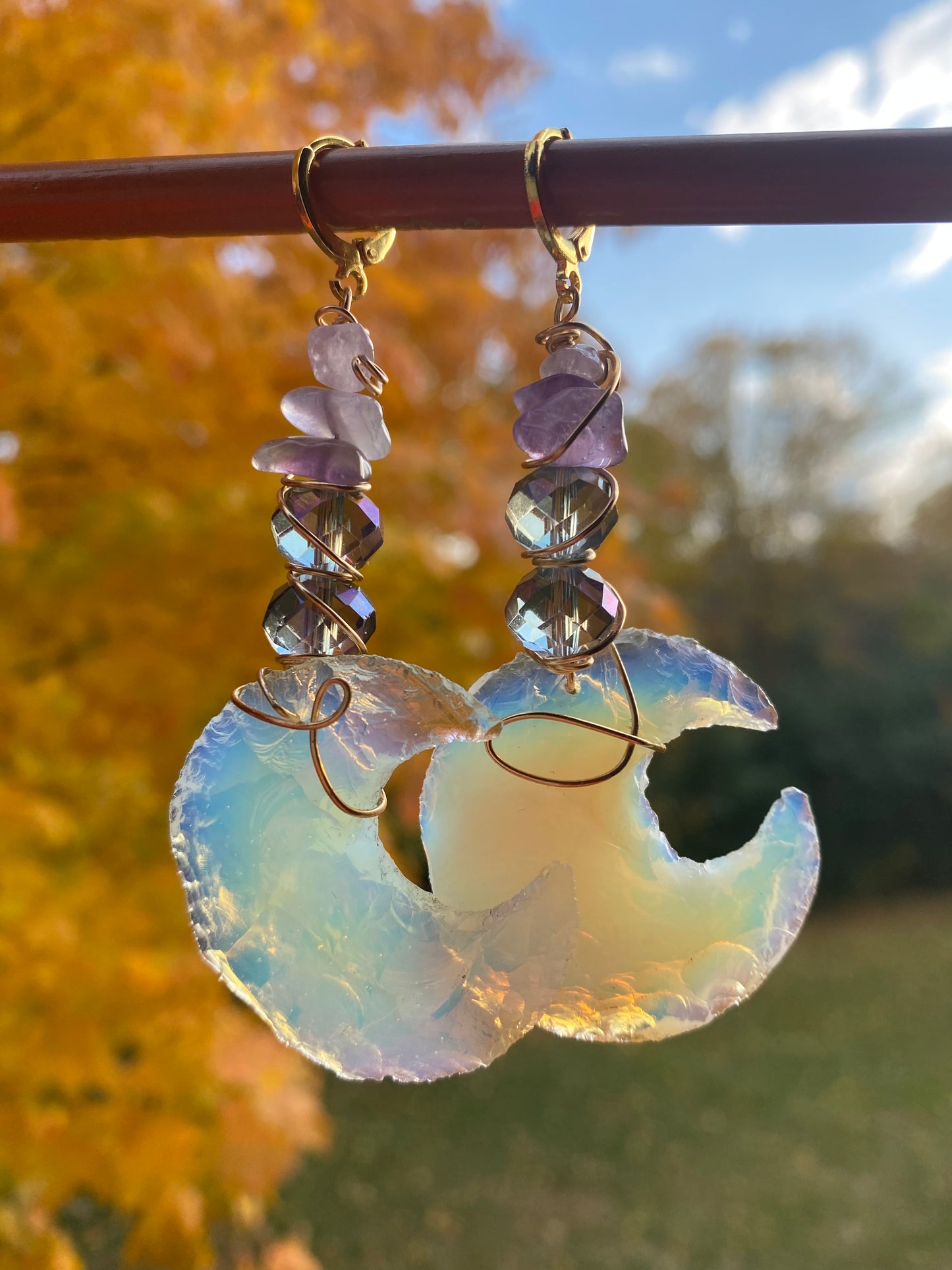 Amethyst and violet iridescent crystal beads with multicolored opalite moons, wrapped with gold wire to create dangle earrings. They are held onto a brown display by gold huggy closures. There is a gold tree and green landscape in the background. 